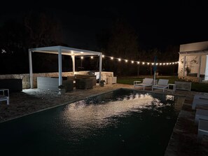 Zona junto a la piscina con un área de descanso cubierta, luces colgantes y un muro de piedra.