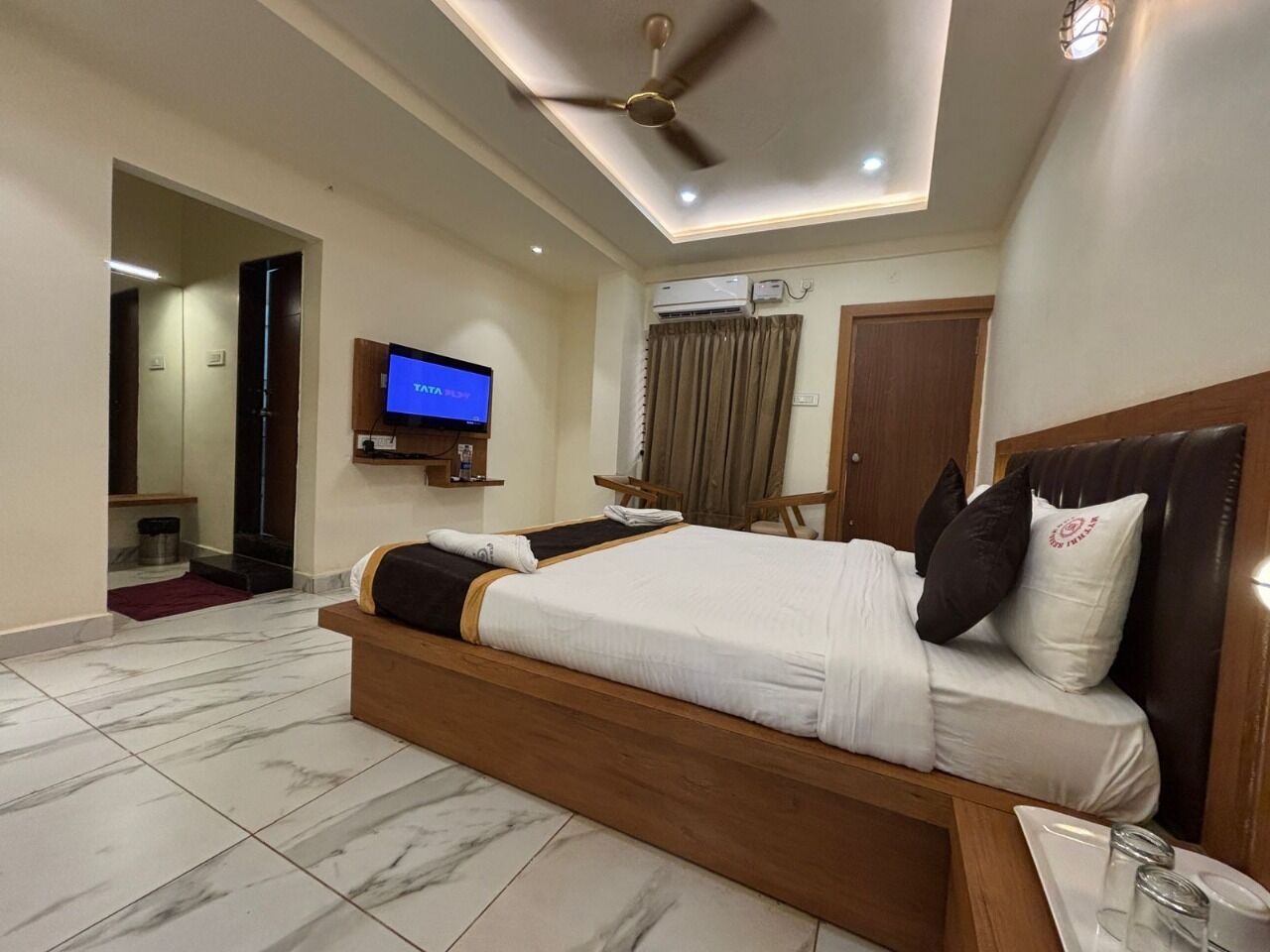 A modern hotel room with a large bed, a ceiling fan, a flat-screen TV, and a wooden headboard.
