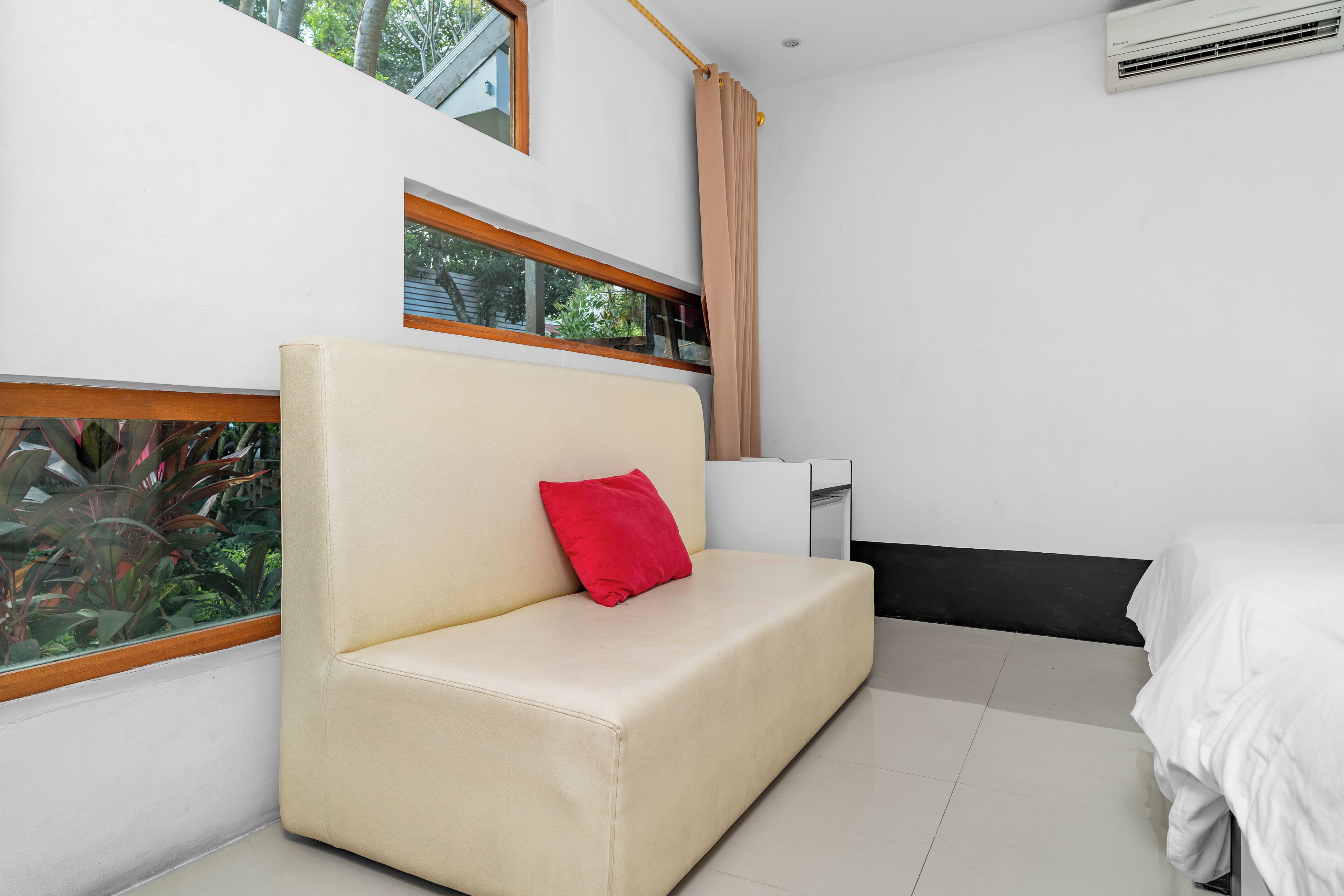 Een moderne kamer met een beige bank, een rood kussen, een wit bed en een raam met uitzicht op groen.