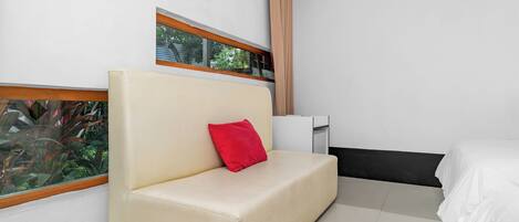 Een moderne kamer met een beige bank, een rood kussen, een wit bed en een raam met uitzicht op groen.