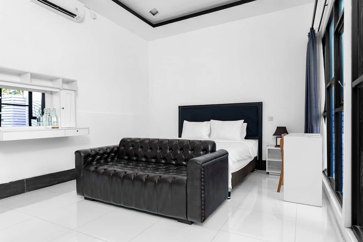 Un dormitorio moderno con un sofá de cuero oscuro, una cama con sábanas blancas, una mesita de noche y un ventanal con cortinas.