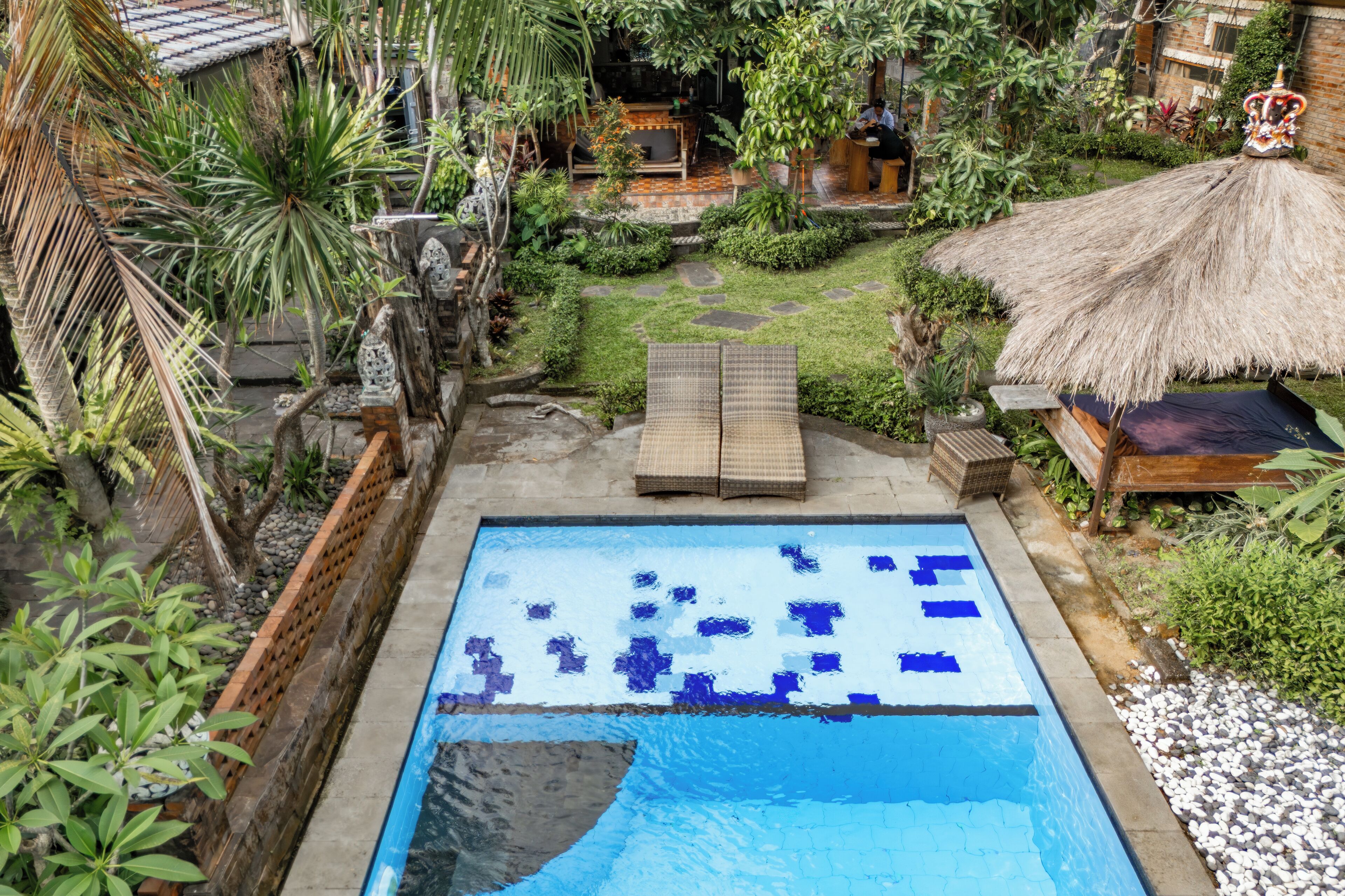 A rectangular swimming pool with a blue and white mosaic design, surrounded by lush greenery and a thatched-roof structure.