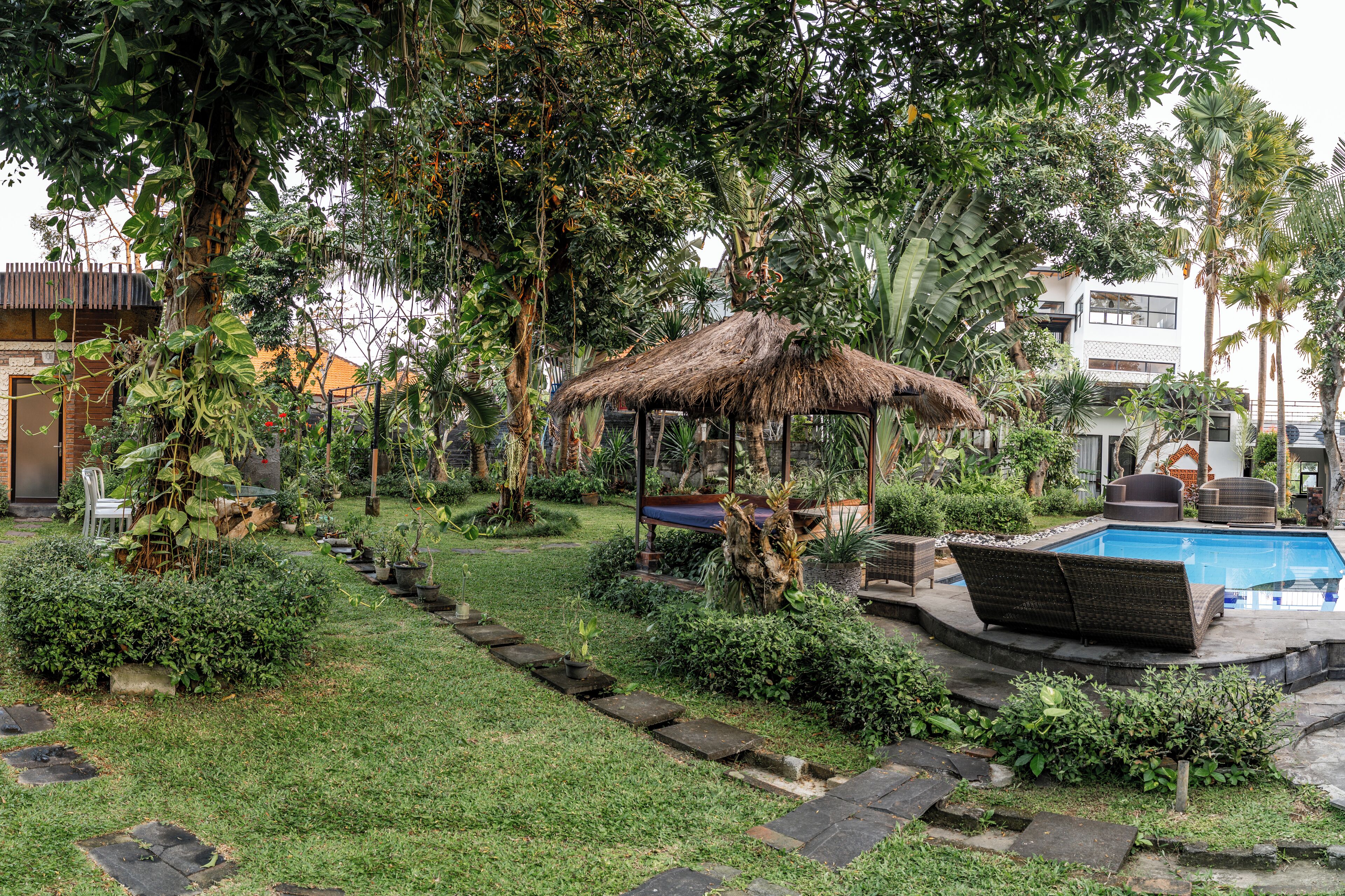 A lush garden with a thatched gazebo, a pool, and various plants.
