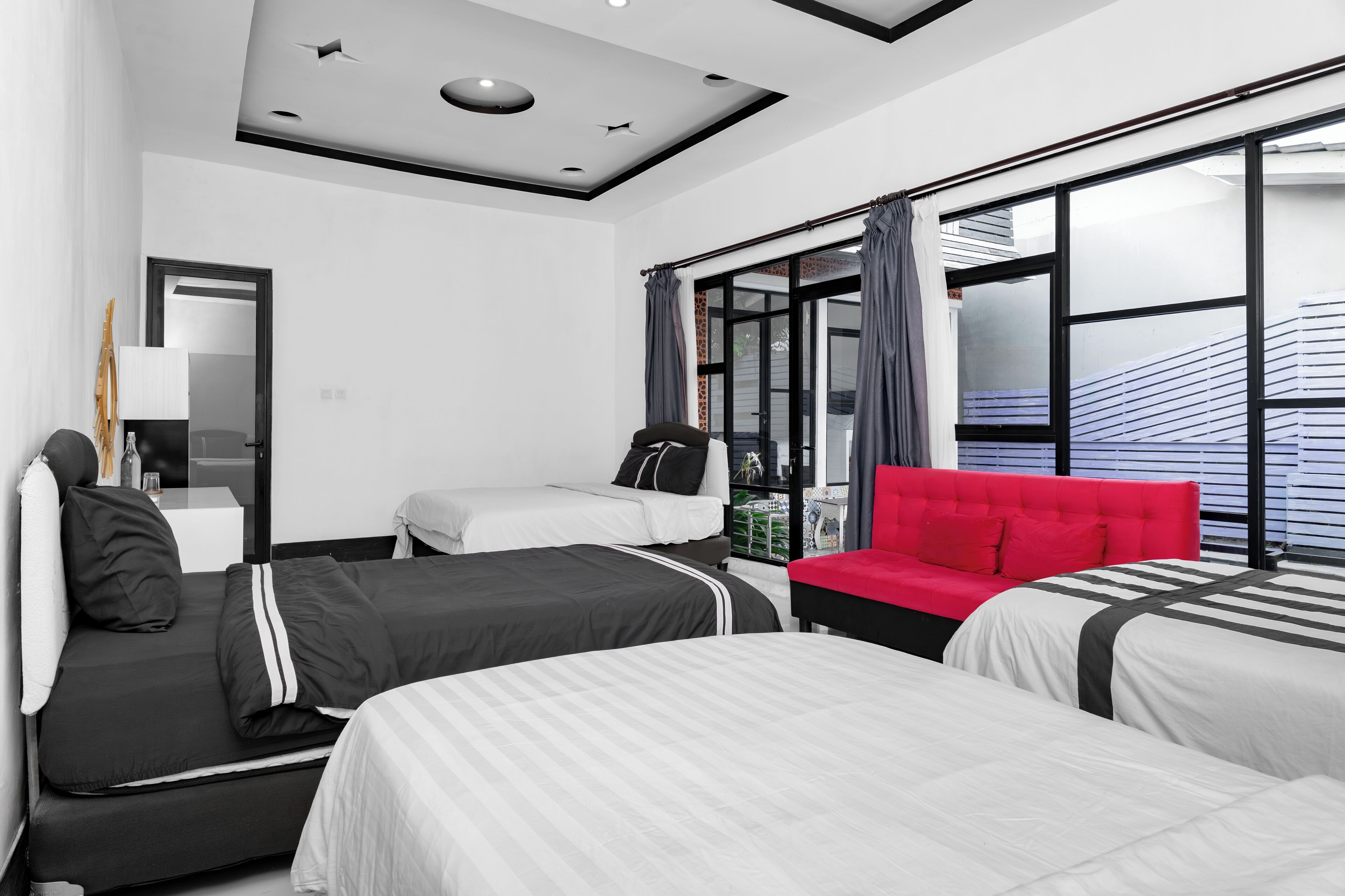 A modern hotel room with three beds, a red sofa, and large windows with blinds.