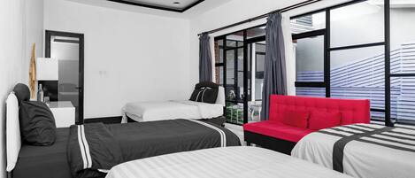 A modern hotel room with three beds, a red sofa, and large windows with blinds.