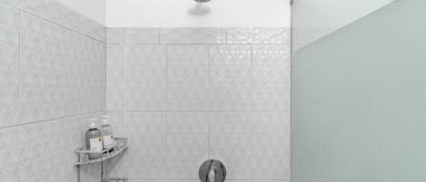Zona de ducha con cabezal de ducha manual, grifo empotrado en la pared y estante con artículos de aseo.
