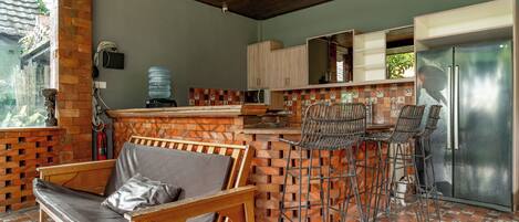 Una cocina con pared de ladrillo, techo de madera, dispensador de agua, refrigerador y una barra con sillas.