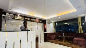 Zona de recepción de un hotel con mostrador de check-in, un letrero que dice "SHREE PALACE", un ventilador y una zona de estar con vistas al exterior.
