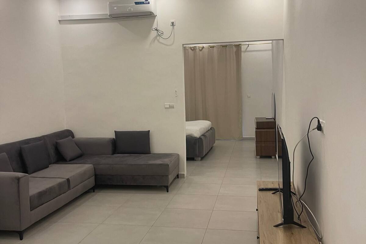 A modern living room with a sectional sofa, a flat-screen TV on a wooden stand, and a ceiling-mounted air conditioner.