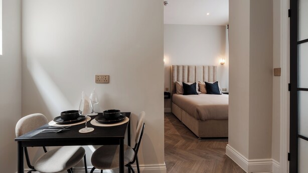 A modern dining area with a table set for two, a sofa in the background, and a wooden floor.