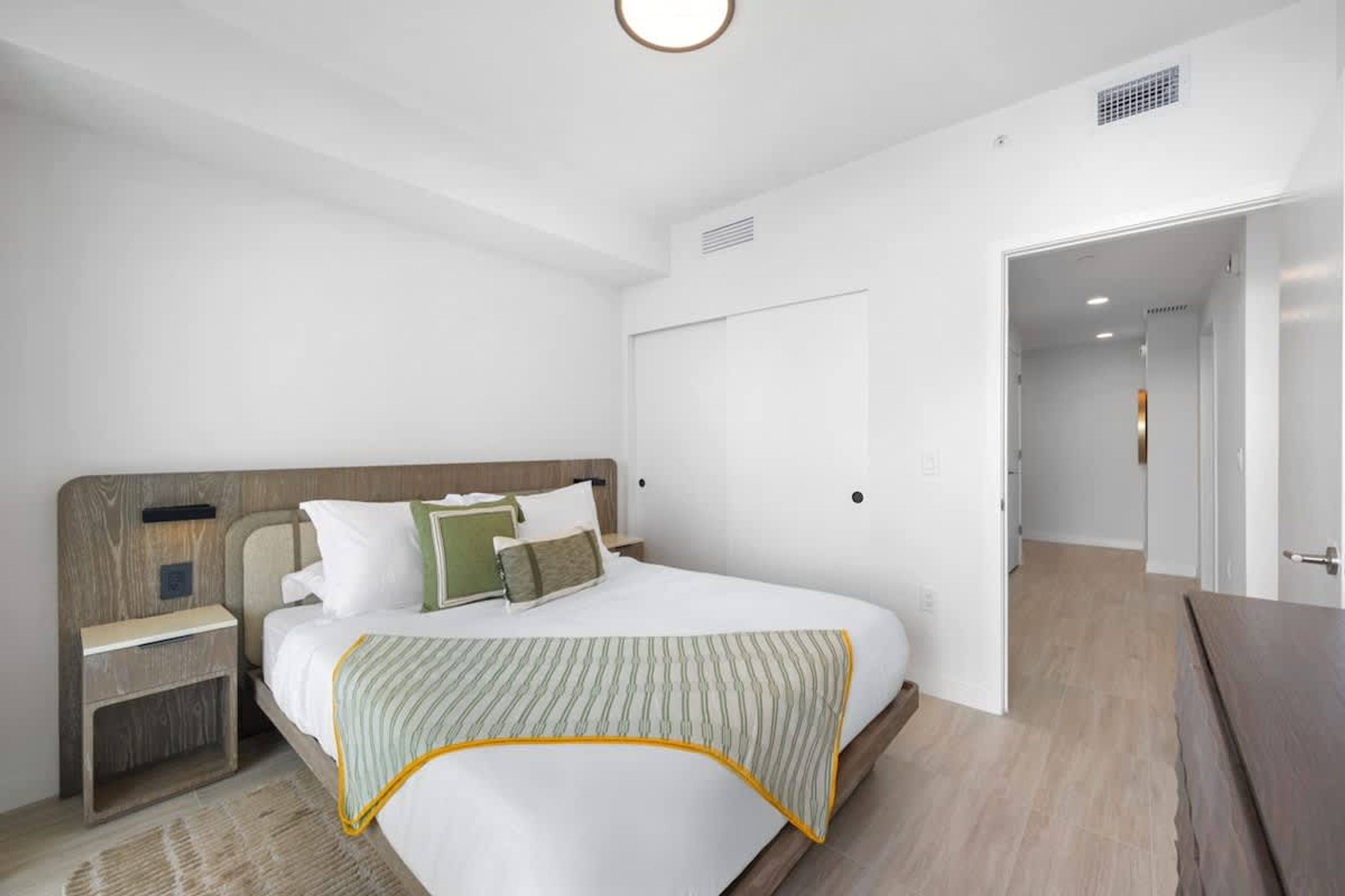 Een moderne hotelkamer met een groot bed, een houten hoofdeinde, een nachtkastje en een deur die naar een andere kamer leidt.