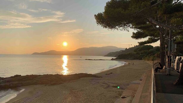 A beach with a sunset, a rocky shoreline, and a paved walkway with benches.