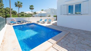 Una piscina con sillones de descanso, una terraza con baldosas de piedra y un edificio blanco con contraventanas azules.