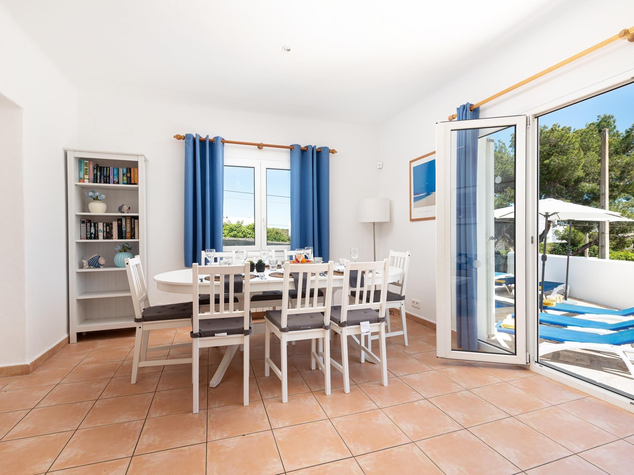 Un comedor con una mesa y sillas blancas, una estantería y vistas al exterior.