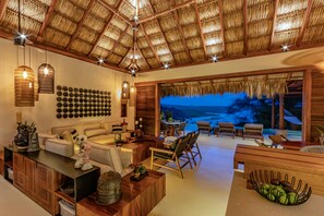 A spacious living area with a thatched ceiling, a wall of round mirrors, and a view of a beach at dusk.