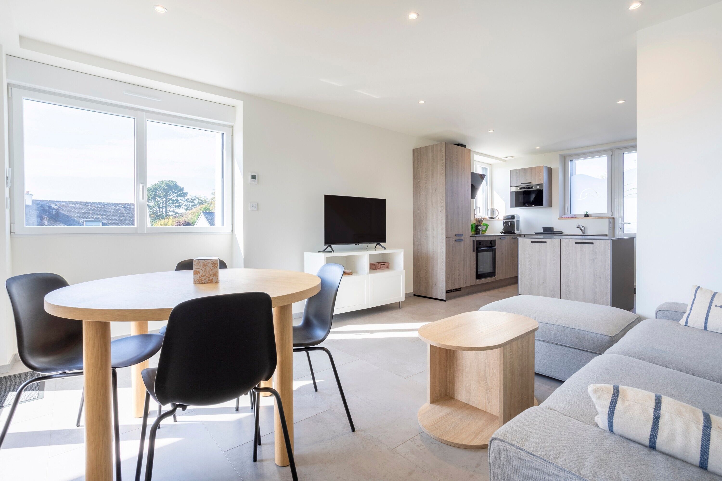 A modern living room with a round dining table, black chairs, a grey sofa, and a small wooden side table.