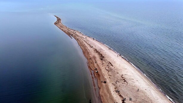 Eine Luftaufnahme eines Strandes mit einer gewundenen Küstenlinie, ruhigem Wasser und vereinzelten Personen am Strand.