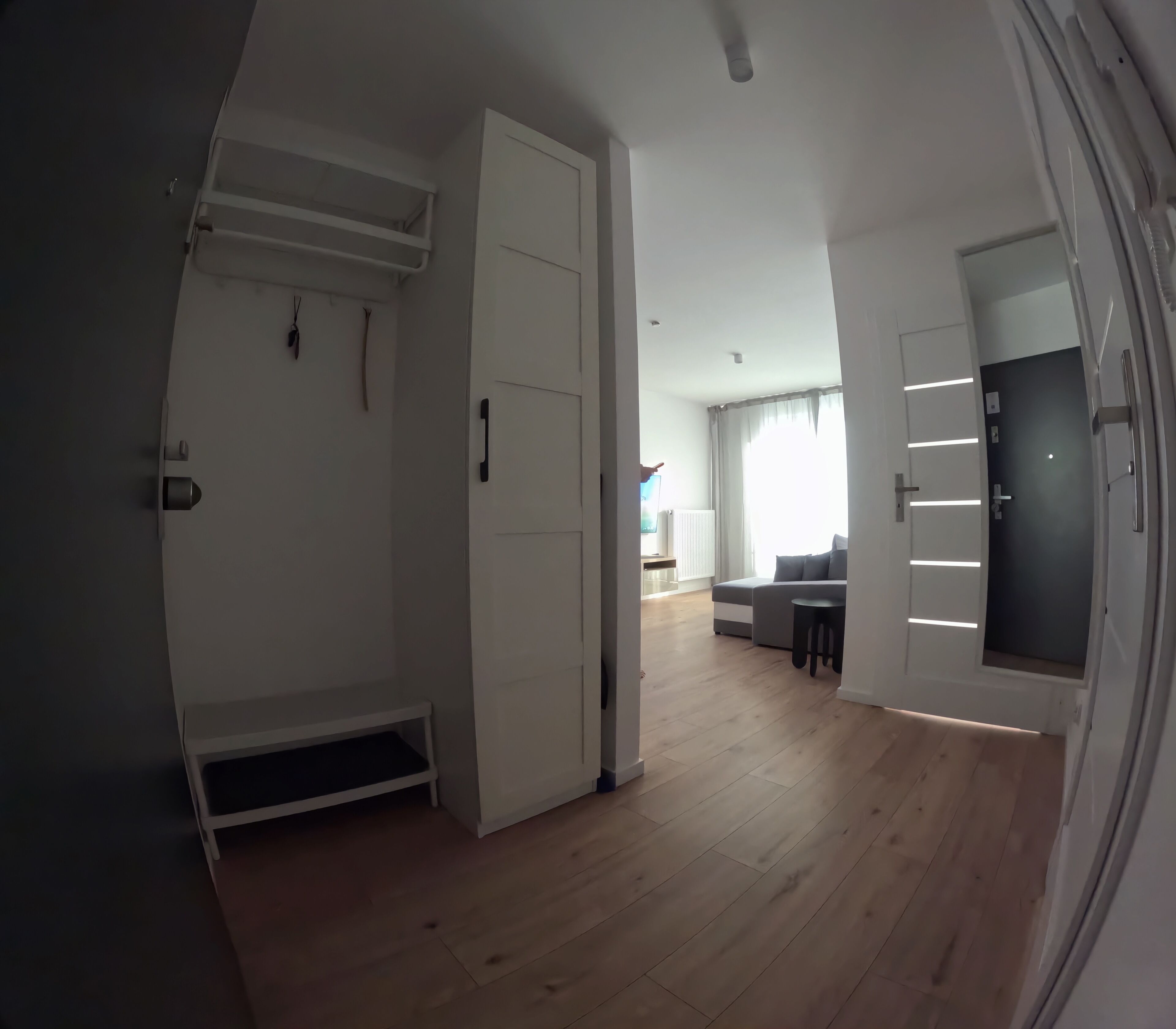 A room with a wooden floor, white walls, a bed, a TV, and a door leading to another room.