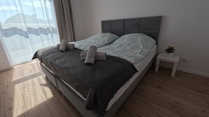 Ein modernes Schlafzimmer mit einem großen Bett, grauer Bettwäsche, einem grauen Kopfteil, einem Nachttisch mit Pflanze und einem Fenster mit Vorhängen.