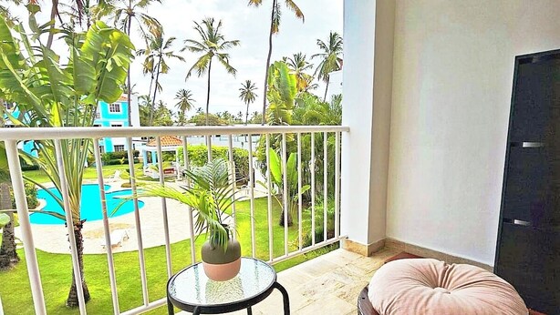 Ein Balkon mit Blick auf einen Swimmingpool, Palmen und ein blaues Gebäude. Zwei runde Tische mit Stühlen und ein Blumentopf sind ebenfalls vorhanden.