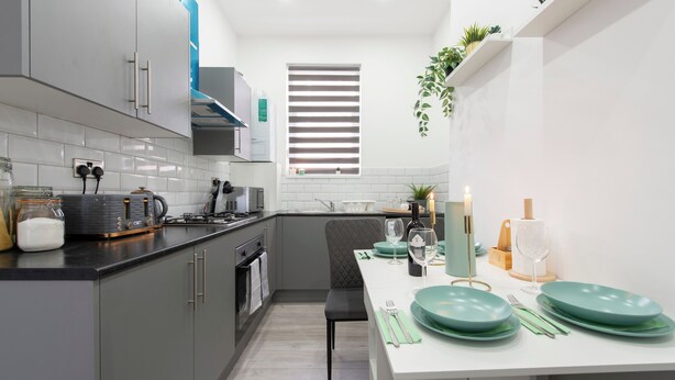 A modern kitchen with grey cabinets, a white countertop, and a dining area set with green plates and glasses. - Apt - Stay 6 - Paid Parking - Petsok - Stratford (London)