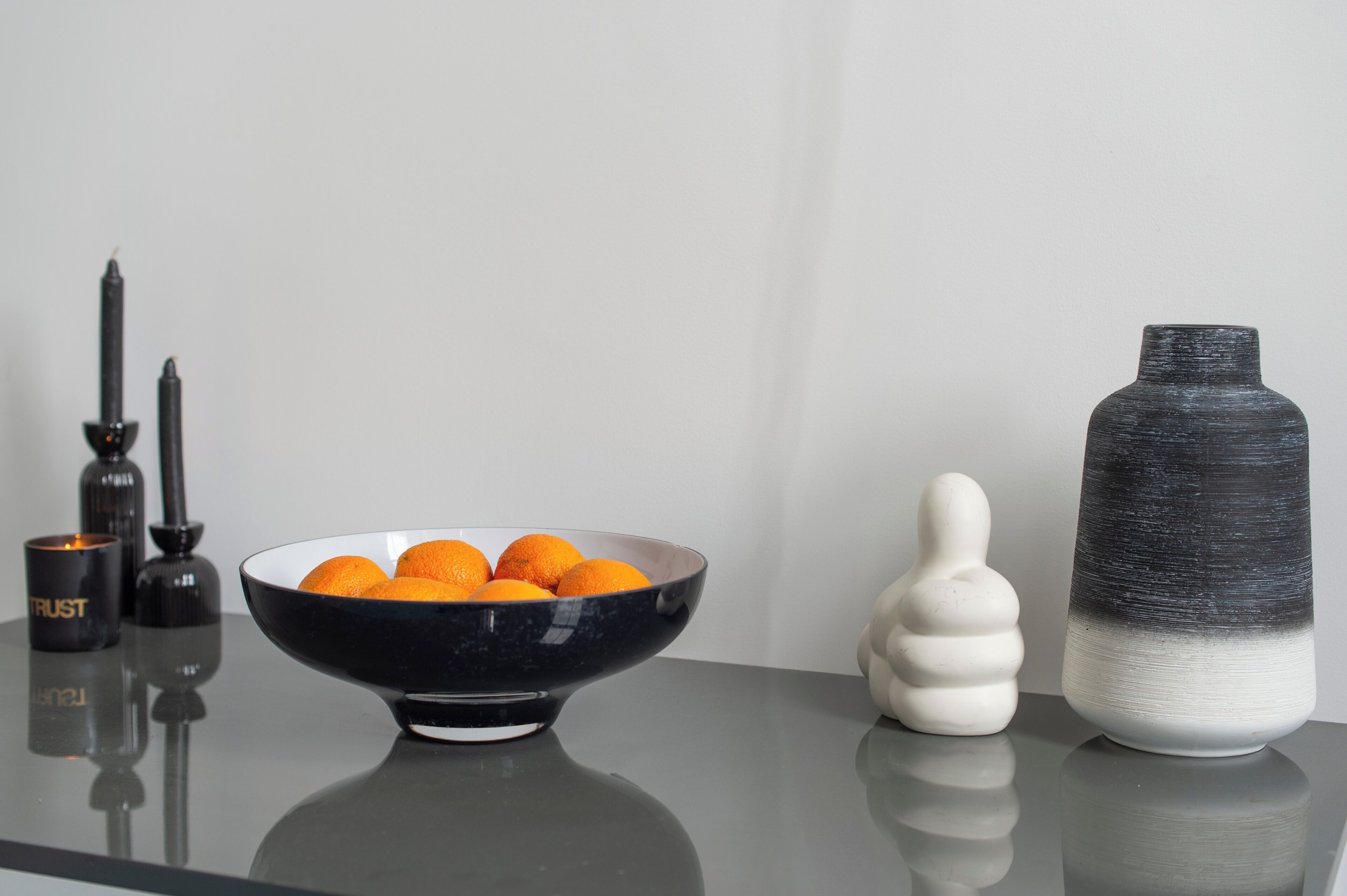 Een zwarte schaal met sinaasappels, een witte sculptuur en een zwart-wit vaas op een reflecterend oppervlak.