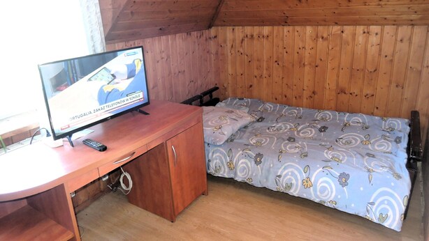 A small room with a wooden ceiling, a bed with a floral bedspread, a desk with a computer and a remote control, and a window with curtains. - Peaceful Stay in Scenic Kolczewo (gmina Wolin)