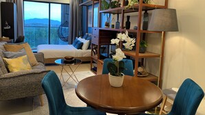 Uma sala de estar moderna com um sofá, uma mesa redonda de madeira e uma poltrona azul. Há uma cama com roupa de cama azul e uma janela grande que oferece vista para o exterior.