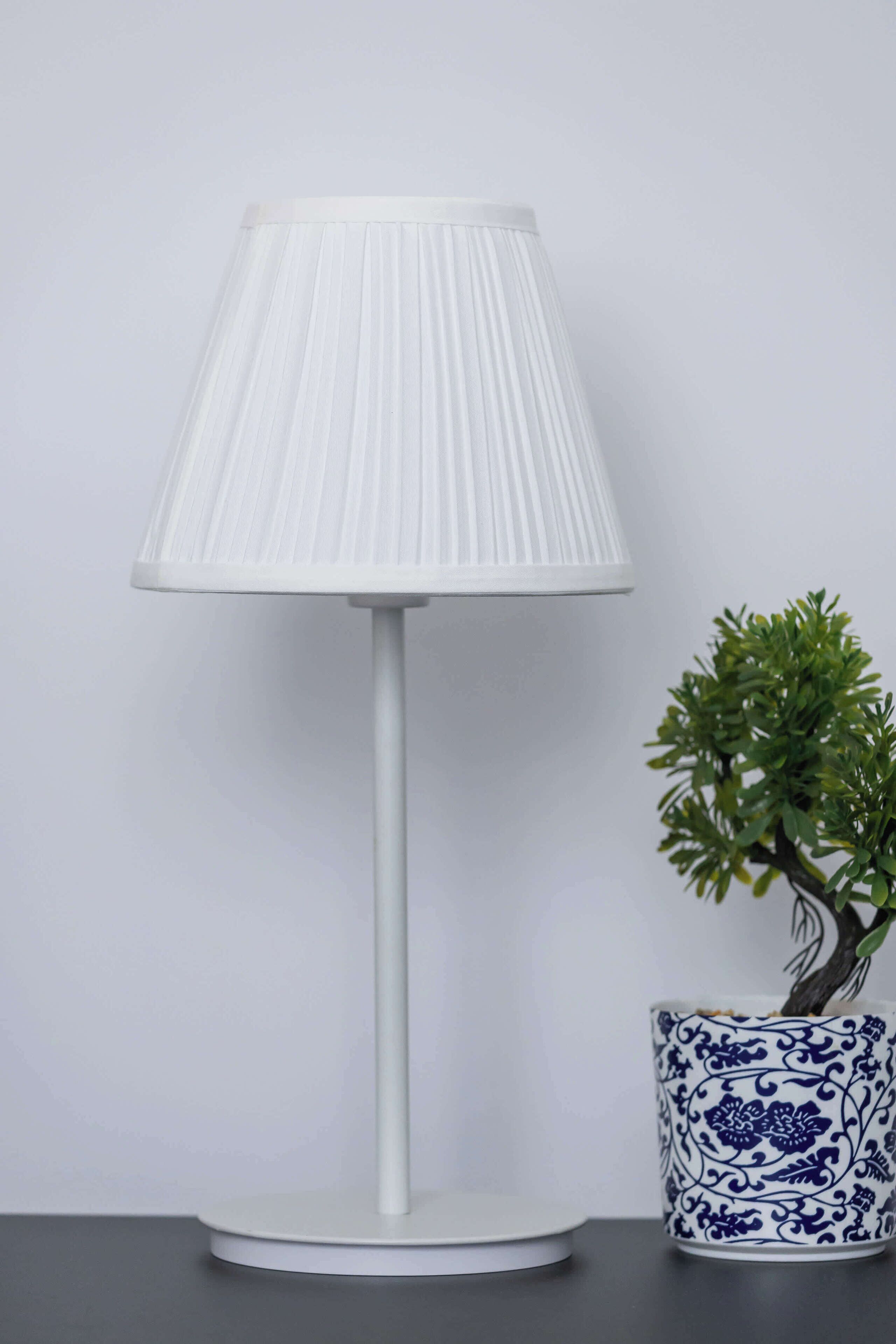 Una lámpara de mesa blanca con pantalla acanalada, una planta en maceta y una jarra de cerámica azul y blanca decorativa.