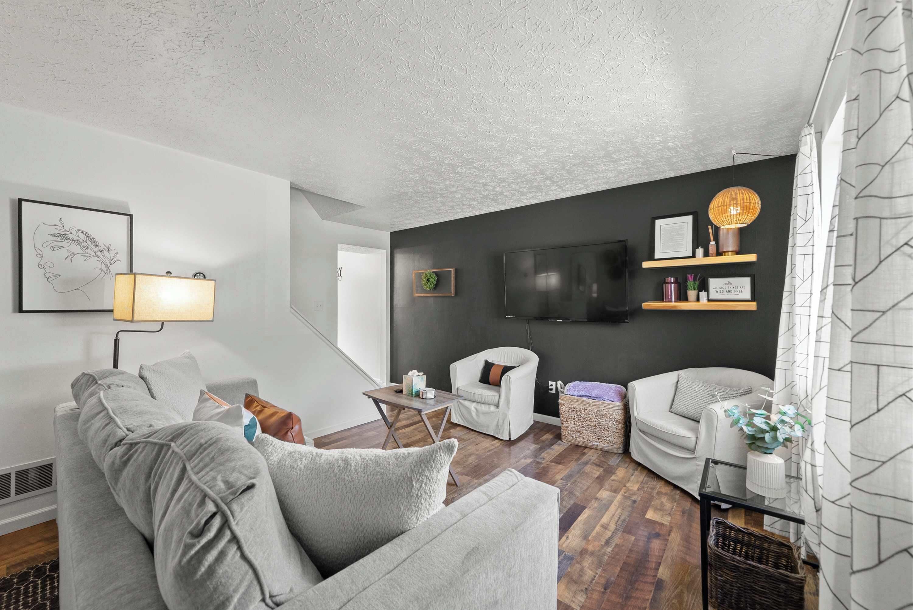 A modern living room with a grey sofa, a white armchair, a small table, a TV mounted on the wall, and a wicker basket.