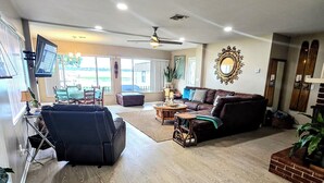 A spacious living room with a large brown leather sofa, a round coffee table, and a wooden floor. There is a television mounted on the wall and a ceiling fan.
