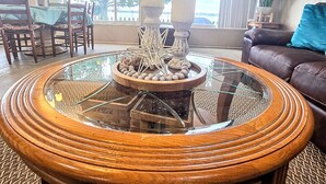 A round wooden coffee table with a glass top and decorative items, a brown leather sofa, and a dining area with a table and chairs.