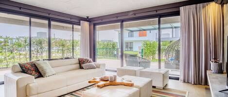 Una sala de estar moderna con un sofá, un reposapiés y una mesa de centro, ventanas amplias y vistas a zonas verdes y edificios.