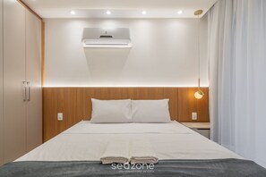 A modern hotel room with a neatly made bed, a wooden headboard, an air conditioning unit, and a bedside lamp. - Apt w/ Pool & BBQ in Bombinhas Rna0303 (Bombinhas)