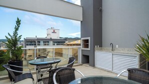 Un balcon avec une table en verre et des chaises en osier, un pot de fleurs et une vue sur un immeuble doté d’une cheminée.