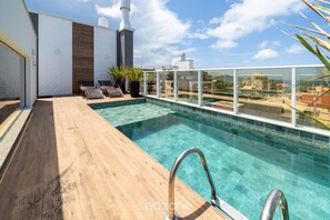 A rooftop pool with clear blue water, a wooden deck, and a view of the cityscape. - Apt w/ Pool & BBQ in Bombinhas Rna0303 (Bombinhas)
