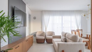Un salon moderne comprenant un canapé, deux fauteuils, un meuble en bois, un téléviseur et une grande fenêtre ornée de rideaux légers.