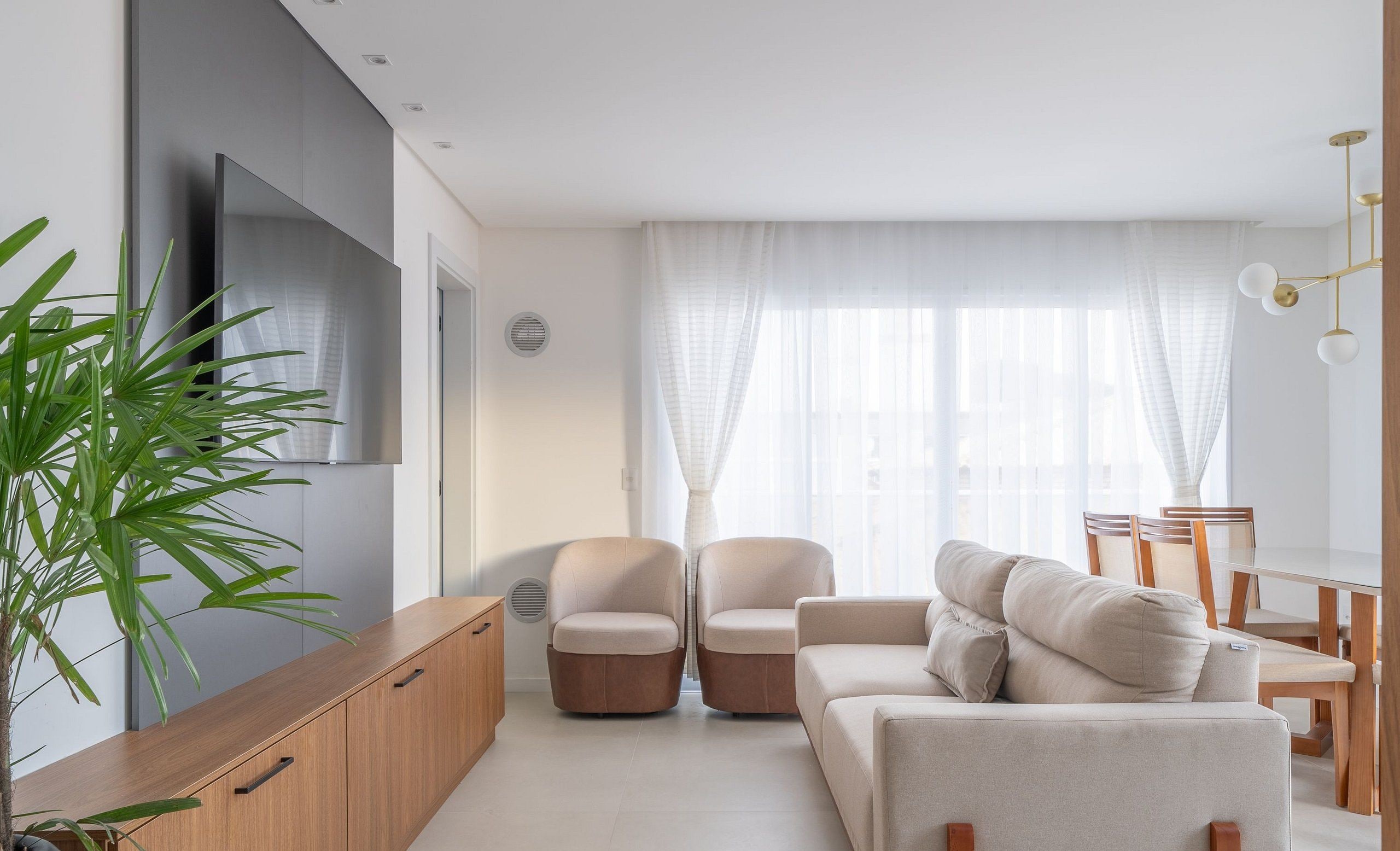 A modern living room with a sofa, two armchairs, a wooden cabinet, a TV, and a large window with sheer curtains.