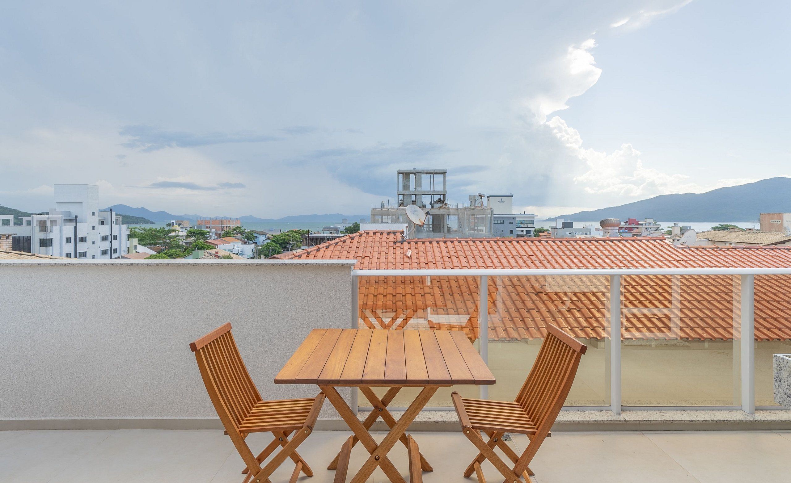 Terraço no telhado com mesa e cadeiras de madeira, com vista para uma paisagem urbana com edifícios e montanhas.
