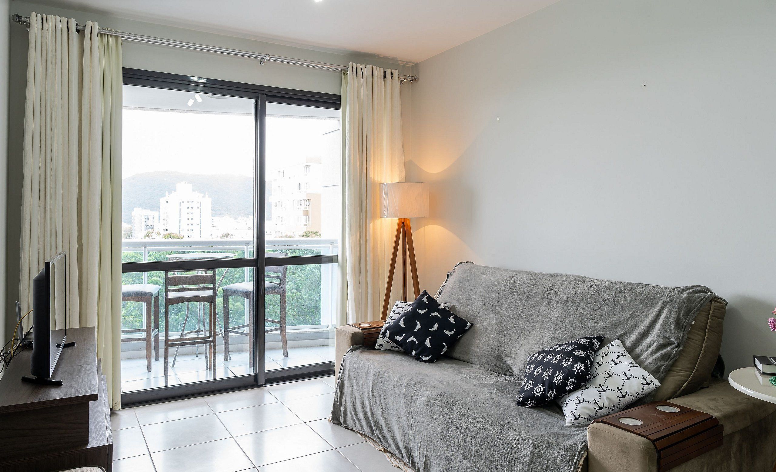 A living room with a grey sofa, a TV on a stand, a lamp, and a view of a balcony with chairs and a table.
