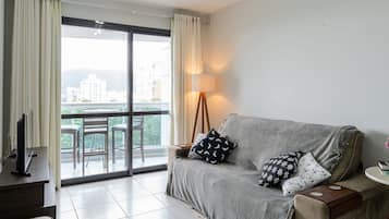 A living room with a grey sofa, a TV on a stand, a lamp, and a view of a balcony with chairs and a table.