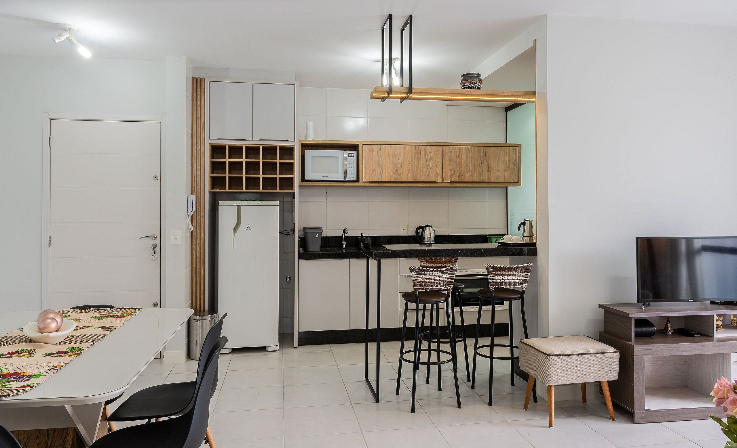 Een moderne keuken met een magnetron, spoelbak en houten keukenkastjes. Er is een eettafel met stoelen en een tv-meubel met een flatscreen televisie.