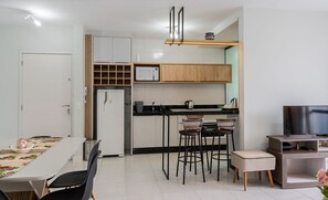 A modern kitchen with a microwave, sink, and wooden cabinets. There is a dining table with chairs and a TV stand with a flat-screen TV. - Apt W/balcony and BBQ 500m From the Beach Rml605 (Imbituba)