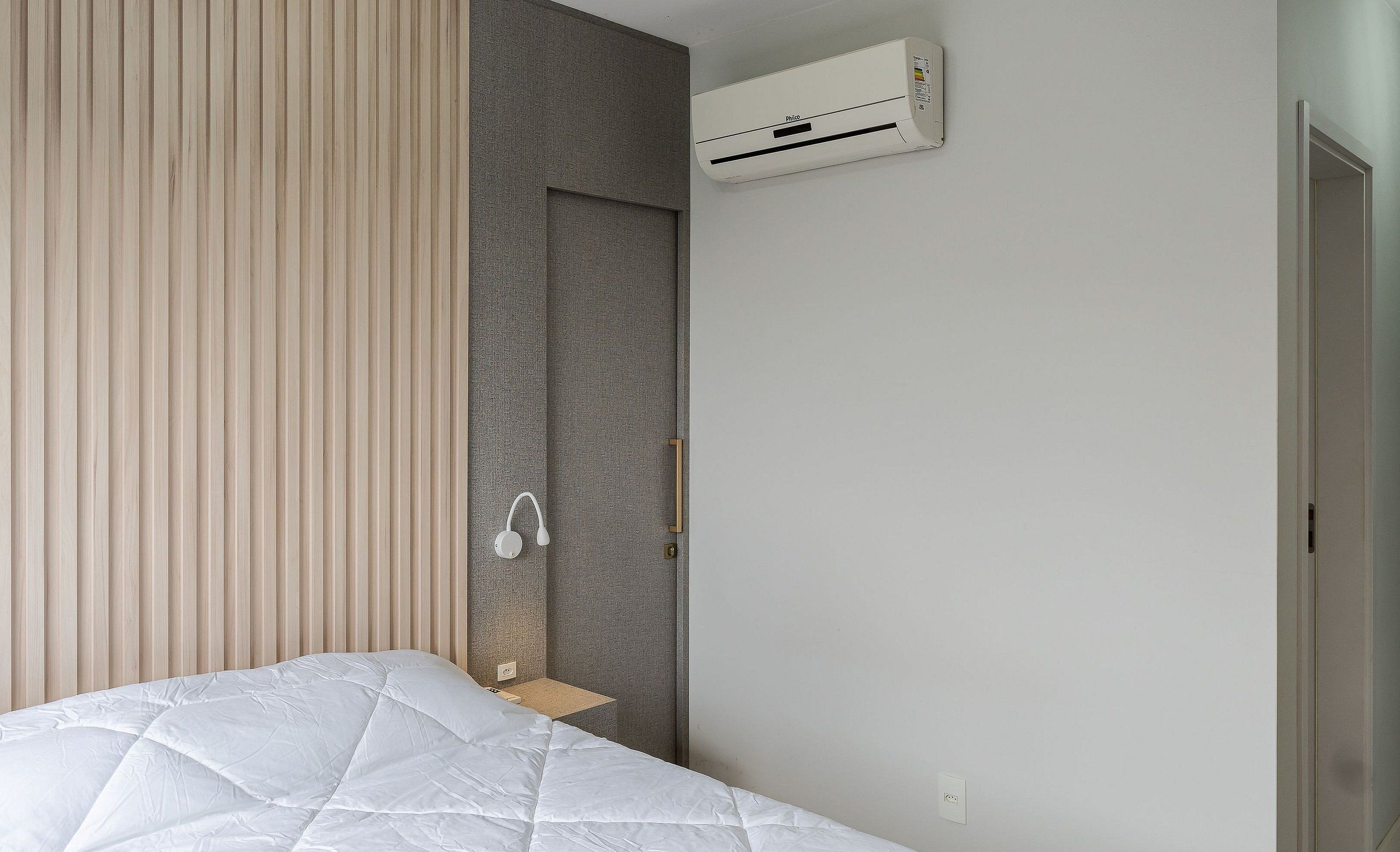 A modern bedroom with a bed, a bedside table, a wall-mounted air conditioner, and a door.