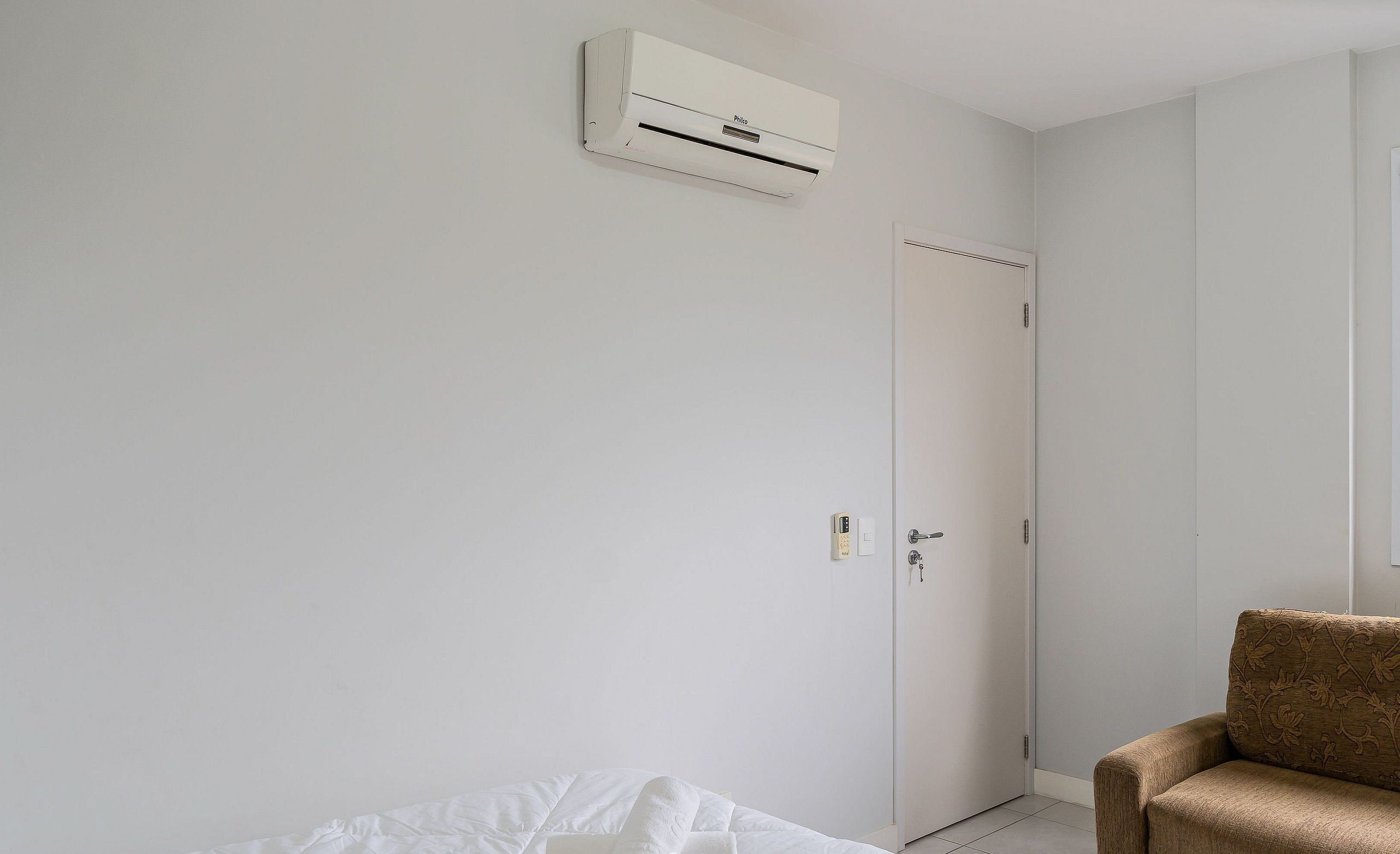 Quarto minimalista com uma cama, uma poltrona marrom, uma porta branca e um ar condicionado fixado na parede.
