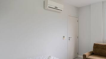 Quarto minimalista com uma cama, uma poltrona marrom, uma porta branca e um ar condicionado fixado na parede.