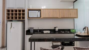 Uma cozinha moderna com micro-ondas integrado, máquina de café e chaleira. Há também uma geladeira, pia e um espaço de refeições com duas cadeiras.