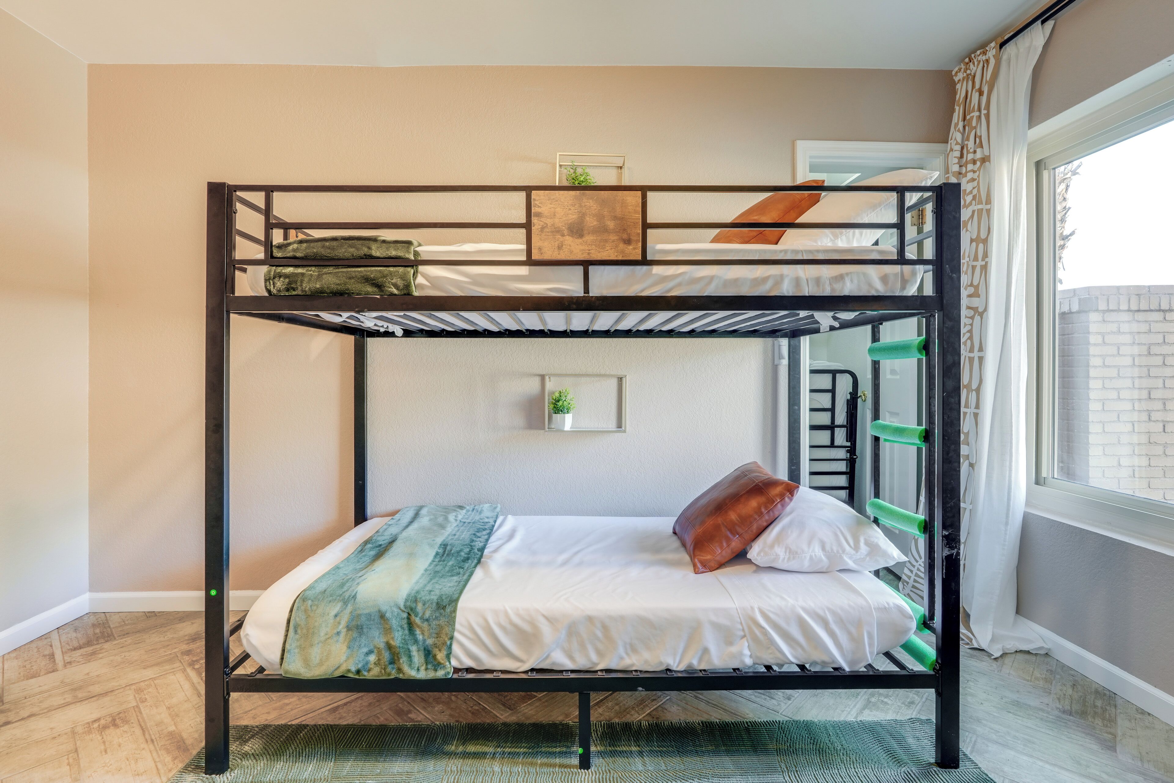 A bunk bed with a green ladder, a small plant on the top bunk, and a window with curtains.