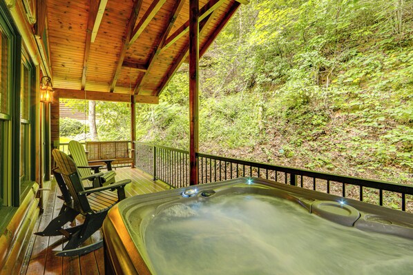 A hot tub on a wooden deck with chairs, surrounded by lush greenery.