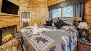 A cozy log cabin room with a fireplace, a bed with a quilt, a mounted TV, and a window with curtains. - 1 Mi to Dtwn Gatlinburg: Hot Tub Haven in Smokies! (Gatlinburg)